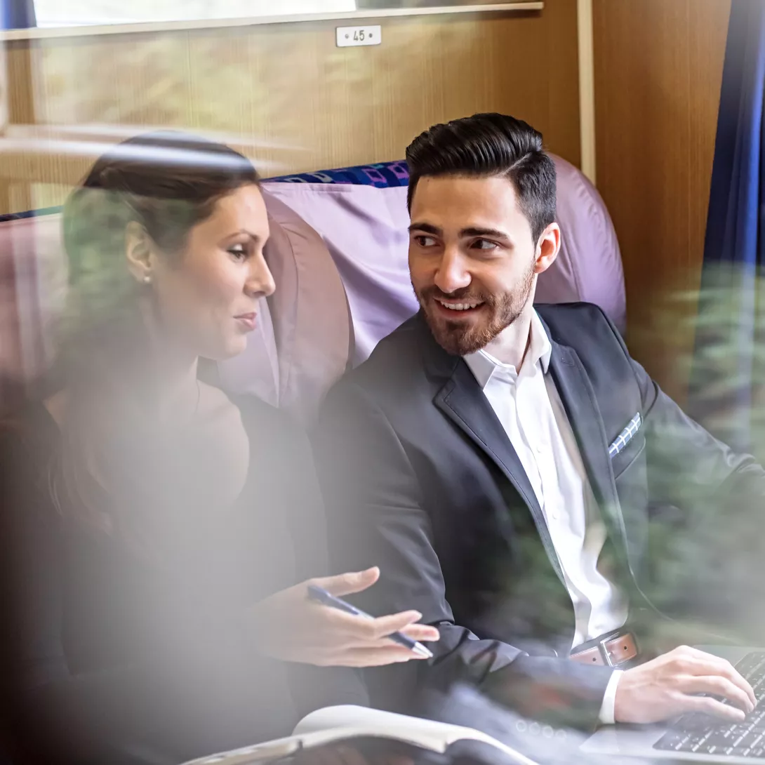 Eine Frau und ein Mann mit Laptop sitzen in einem Zugabteil und diskutieren