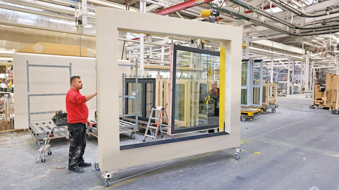 Weiter geht es in der stehenden Wandfertigung der Fabrikstraße: Die Produktionsmitarbeiter Johannes Schmid und Kire Mirchovski (von links) bestücken das fertige Wandmodul mit einem festverglasten Fenster. Neben den Fenstern werden in diesem Produktionsbereich auch Haustüren sowie Rollläden direkt montiert.